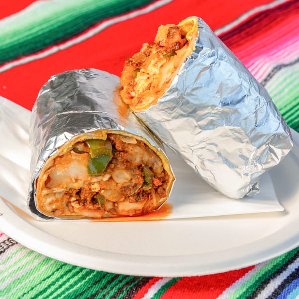 Two foil-wrapped burritos, one cut open to show savory filling, on a white plate with a colorful Mexican blanket.