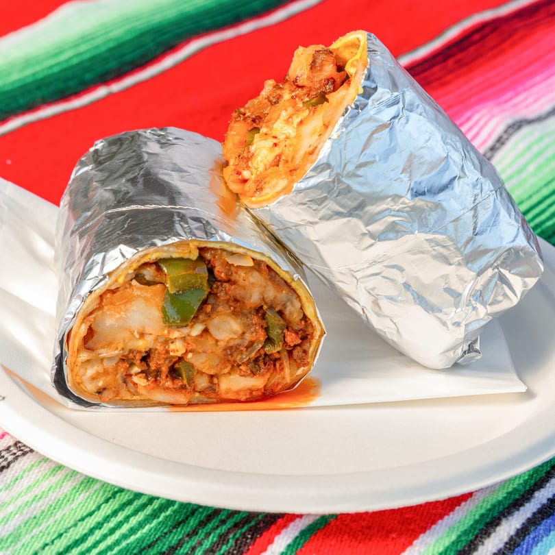 Two foil-wrapped burritos, one cut open to show savory filling, on a white plate with a colorful Mexican blanket.
