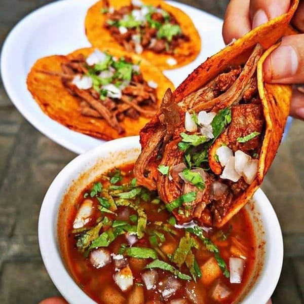 A hand holds a birria taco over a bowl of consommé, with two more tacos in the background.