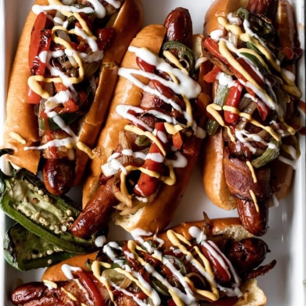 Four bacon-wrapped hot dogs in buns, topped with grilled peppers, onions, mustard, and crema.