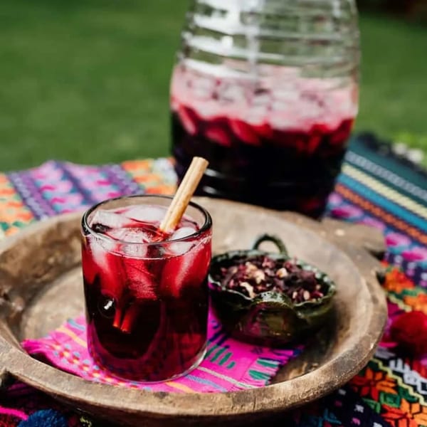 A glass of red hibiscus agua fresca with a cinnamon stick, a jar, and dried hibiscus flowers.