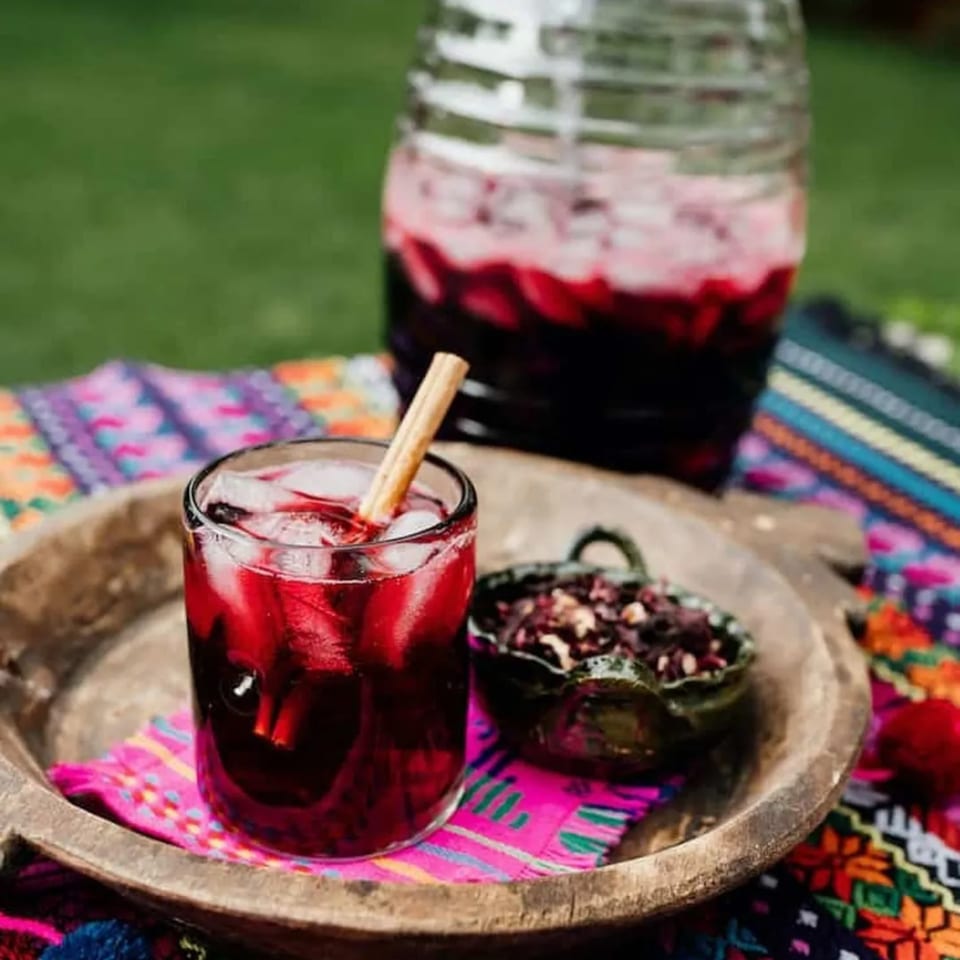 A glass of red hibiscus agua fresca with a cinnamon stick, a jar, and dried hibiscus flowers.