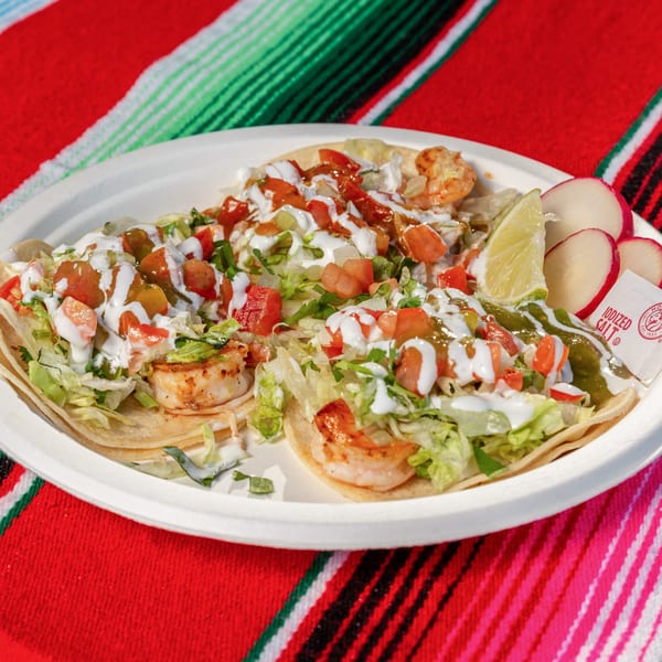 Three shrimp tacos with lettuce, tomatoes, and sauces, served with lime and radishes on a Mexican blanket.