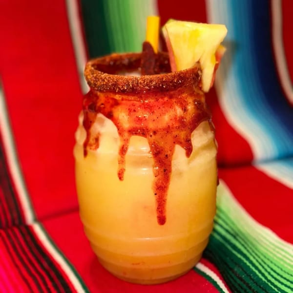 A vibrant yellow Mexican drink with chili-lime rim, red chamoy drizzle, and pineapple garnish.